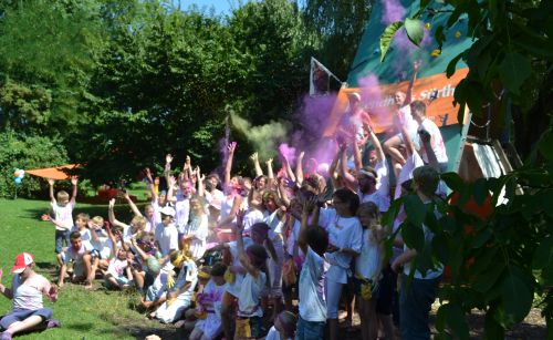 Teilnehmer/innen einer Ferienaktion des Jugendhauses Sürth in weißen Shirts reißen jubelnd die Arme in die Höhe und werfen dabei lilafarbenes Pulver in die Luft