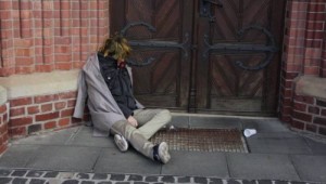 Screenshot aus dem Film Dasein: Ein zusammengesunkener Bettler vor einem Kircheneingang