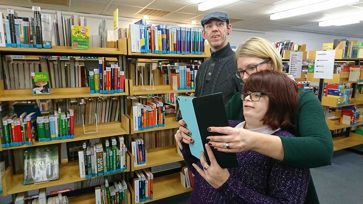 Frau fotografiert mit Tablet in Bibliothek, sie wird unterstützt von einer Assistenz. Ein Mann schaut in die Kamera