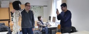 Junge fotografiert lachenden Jungen, der ein Skelett mit einer Fellmütze in einem Klassenzimmer umarmt. Im Hintergrund ein weiterer Junge am PC