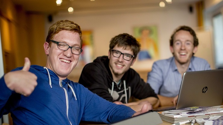 3 junge Männe mit und ohne Lernschwierigkeiten lächelnd vor Laptop, einer zeigt Daumen hoch.