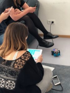 Frau sitzt auf Boden und hält Tablet, links zwei Personen auf Couch, auf Boden ist kleiner Roboter