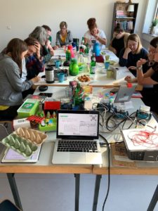 langer Tisch mit Essen, Trinken und Technik, jeweils zwei Personen sitzen zusammen mit Stift und Papier