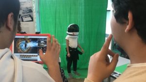 Zwei Jugendliche fotografieren mit der Greenscreen-App ein Kind mit Raumfahrerhelm vor einem grünen Hintergrund