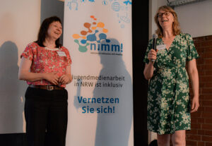 Dr. Christine Ketzer und Susanne Böhmig auf der Bühne mit einem Mikrophon. Hinter ihnen ein Aufsteller mit der Aufschrift nimm! Netzwerk Inklusion mit Medien. Vernetzen Sie sich!