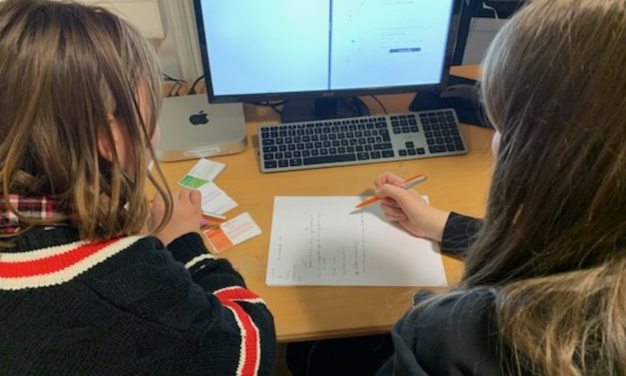 Zwei Personen sitzen an einem PC und schreiben etwas auf ein Blatt Papier