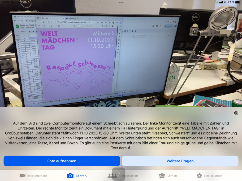 Screenshot der App Be My Eyes: Fotoszene mit Beschreibungstext: 
Auf dem Bild sind zwei Computermonitore auf einem Schreibtisch zu sehen. Der linke Monitor zeigt eine Tabelle mit Zahlen und Uhrzeiten. Der rechte Monitor zeigt ein Dokument mit einem lila Hintergrund und der Aufschrift "WELT MÄDCHEN TAG" in Großbuchstaben. Darunter steht "Mittwoch 11.10.2023 15-20 Uhr". Weiter unten steht "Respekt, Schwester!" und es gibt eine Zeichnung von zwei Händen, die sich die kleinen Finger verschränken. Auf dem Schreibtisch befinden sich auch verschiedene Gegenstände wie Visitenkarten, eine Tasse, Kabel und Boxen. Es gibt auch eine Postkarte mit dem Bild einer Frau und einige grüne und gelbe Kästchen mit Text darauf.