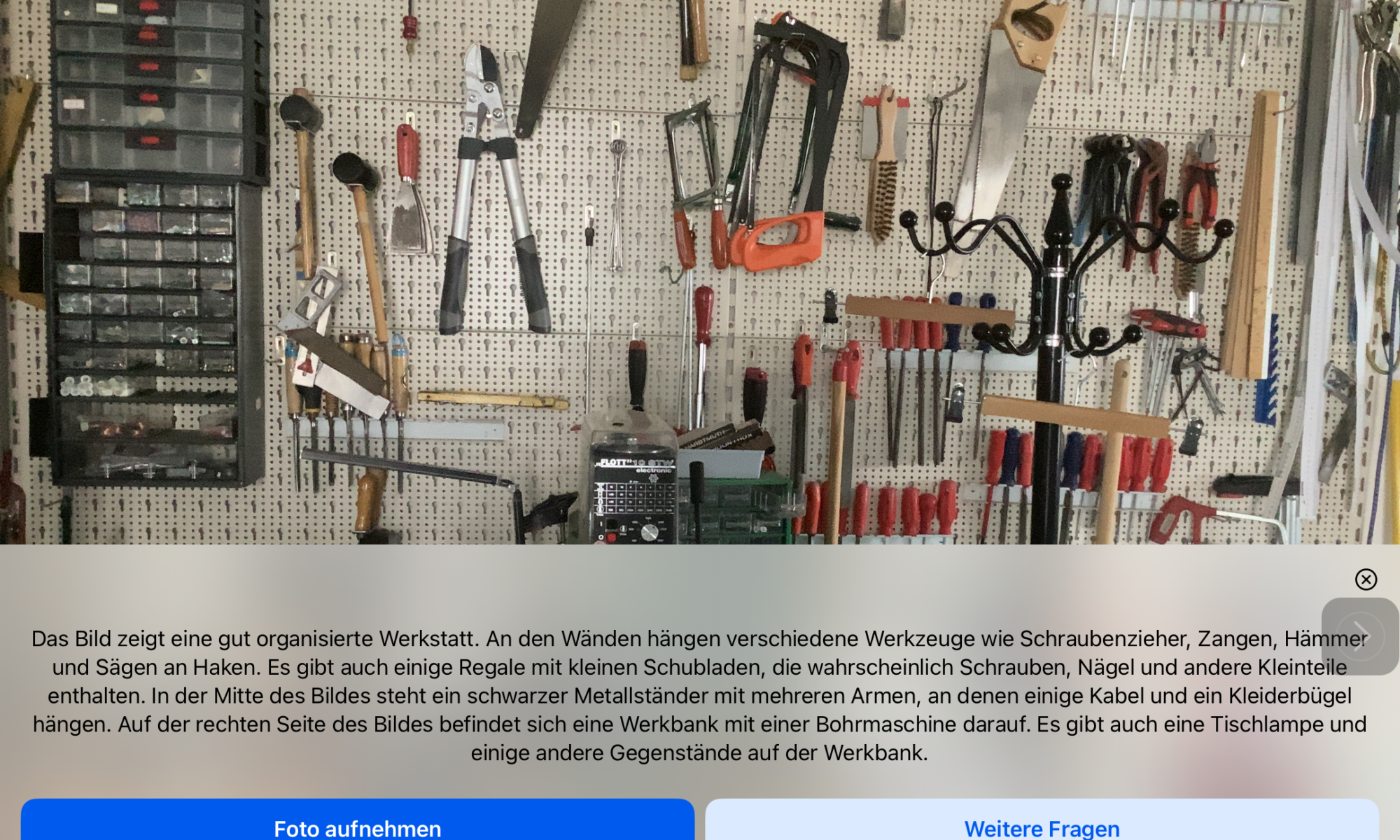 Screenshot der App Be My Eyes: Fotoszene mit Beschreibungstext: Das Bild zeigt eine gut organisierte Werkstatt. An den Wänden hängen verschiedene Werkzeuge wie Schraubenzieher, Zangen, Hämmer und Sägen an Haken. Es gibt auch einige Regale mit kleinen Schubladen, die wahrscheinlich Schrauben, Nägel und andere Kleinteile enthalten. In der Mitte des Bildes steht ein schwarzer Metallständer mit mehreren Armen, an denen einige Kabel und ein Kleiderbügel hängen. Auf der rechten Seite des Bildes befindet sich eine Werkbank mit einer Bohrmaschine darauf. Es gibt auch eine Tischlampe und einige andere Gegenstände auf der Werkbank.