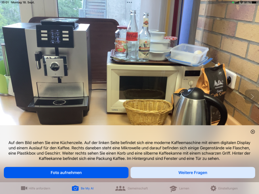 Screenshot der App Be My Eyes: Fotoszene mit Beschreibungstext: Auf dem Bild sehen Sie eine Küchenzeile. Auf der linken Seite befindet sich eine moderne Kaffeemaschine mit einem digitalen Display und einem Auslauf für den Kaffee. Rechts daneben steht eine Mikrowelle und darauf befinden sich einige Gegenstände wie Flaschen, eine Plastikbox und Geschirr. Weiter rechts sehen Sie einen Korb und eine silberne Kaffeekanne mit einem schwarzen Griff. Hinter der Kaffeekanne befindet sich eine Packung Kaffee. Im Hintergrund sind Fenster und eine Tür zu sehen.