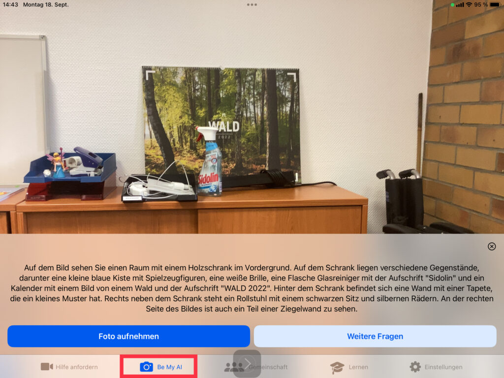 Screenshot der App Be My Eyes: Fotoszene mit Beschreibungstext: Auf dem Bild sehen Sie einen Raum mit einem Holzschrank im Vordergrund. Auf dem Schrank liegen verschiedene Gegenstände, darunter eine kleine blaue Kiste mit Spielzeugfiguren, eine weiße Brille, eine Flasche Glasreiniger mit der Aufschrift "Sidolin" und ein Kalender mit einem Bild von einem Wald und der Aufschrift "WALD 2022". Hinter dem Schrank befindet sich eine Wand mit einer Tapete, die ein kleines Muster hat. Rechts neben dem Schrank steht ein Rollstuhl mit einem schwarzen Sitz und silbernen Rädern. An der rechten Seite des Bildes ist auch ein Teil einer Ziegelwand zu sehen.