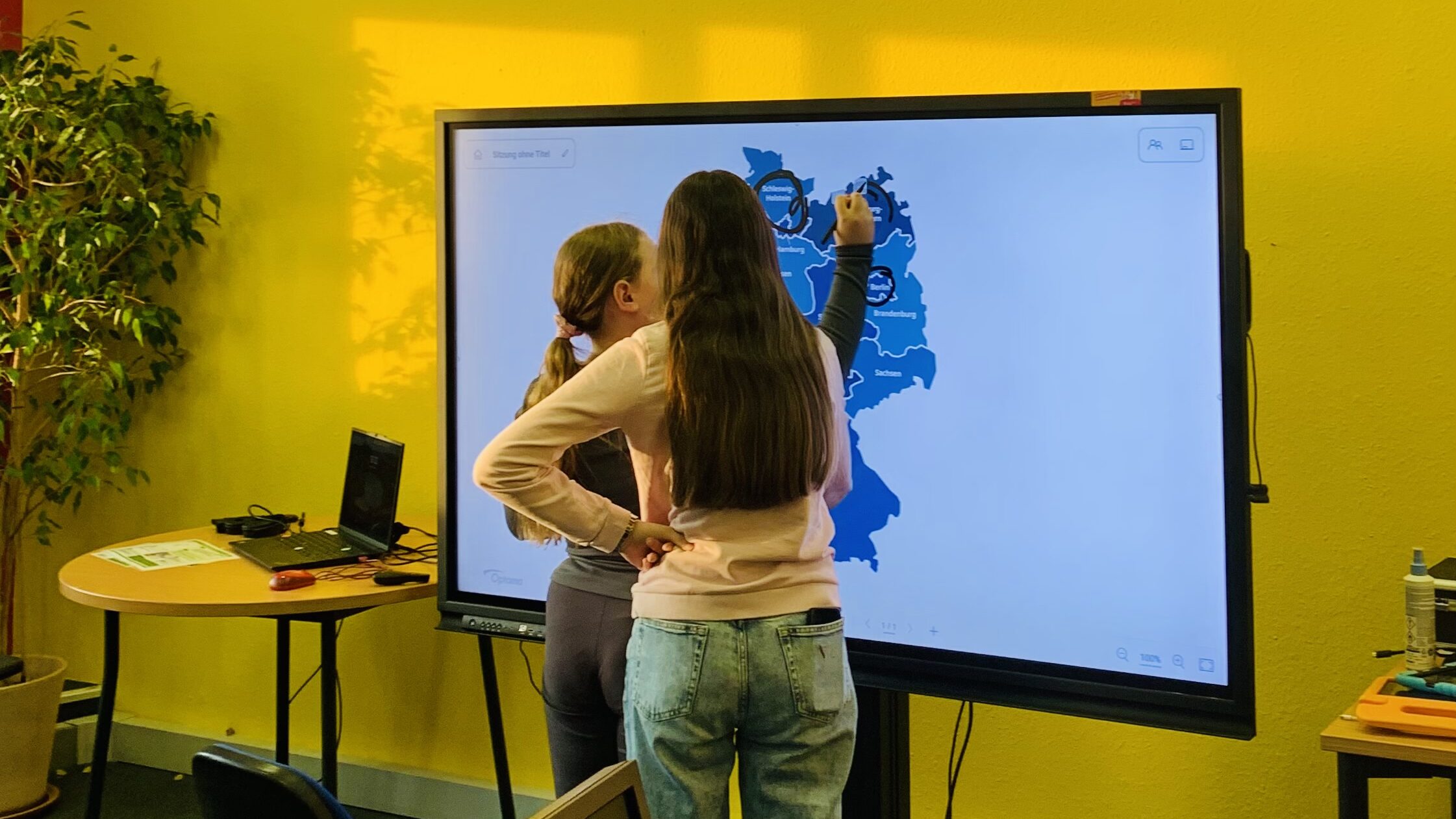 Zwei Mädchen stehen vor einem SmartScreen und kreisen Städte auf einer blauen Landkarte ein.