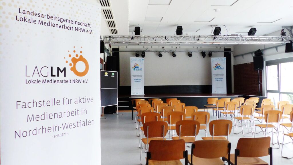 Der leere Saal voller Stühle, im Vordergrund ein Banner der LAG Lokale Medienarbeit. Im Hintergrund die Bühne mit Bannern von nimm!