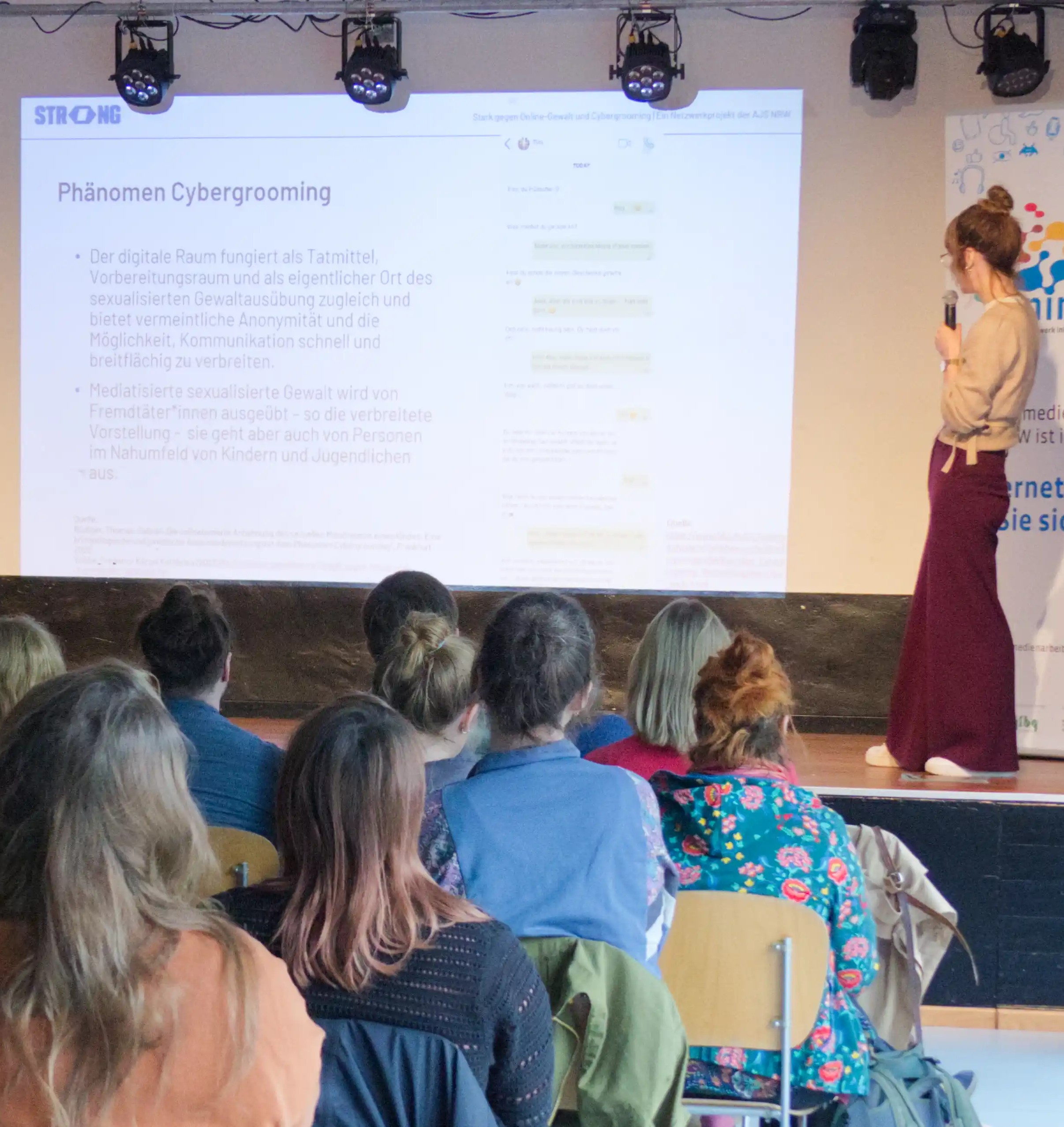 Die Referentin steht vor dem Publikum auf der Bühne. Der Beamer wirft einen Text an: "Phänomen Cybergrooming"