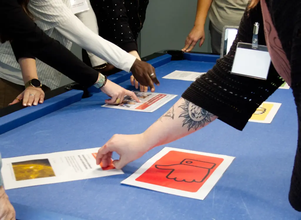 Teilnehmende greifen nach Beispielbildern, die auf einem Tisch ausliegen und ordnen sie.
