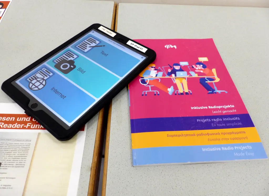 Ein Tablet mit einer Barrierefreiheits-App geöffnet. Daneben ein Flyer der tjfbg zu barrierefreien Radiosendungen.