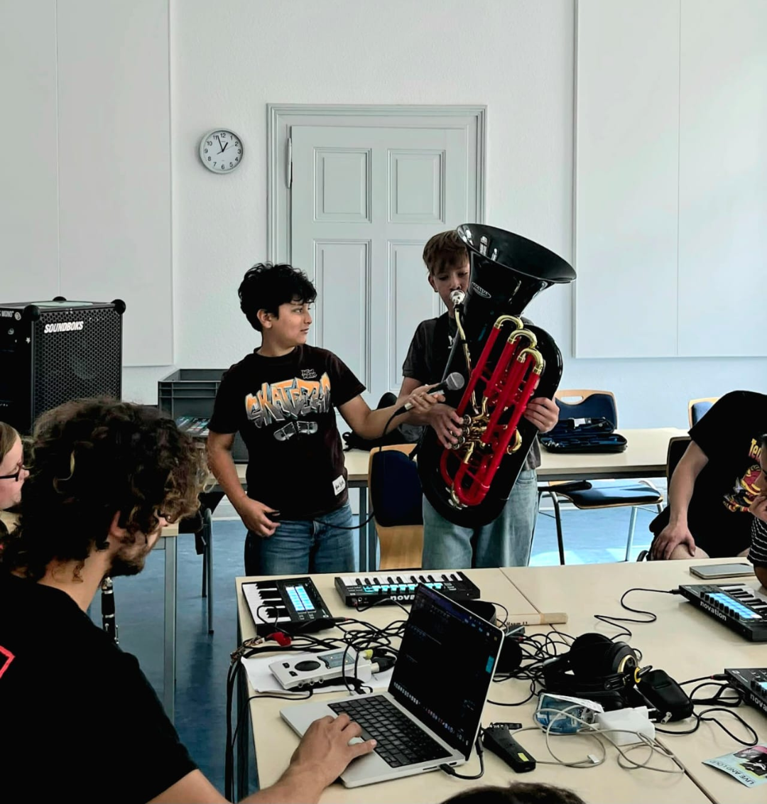 Zwei Jungen stehen in einem Raum, einer hält eine schwarze Tube, der andere Junge hält ein Mikrofon. Mehrere Personen sitzen an einem Tisch mit Laptops und Musikequipment.