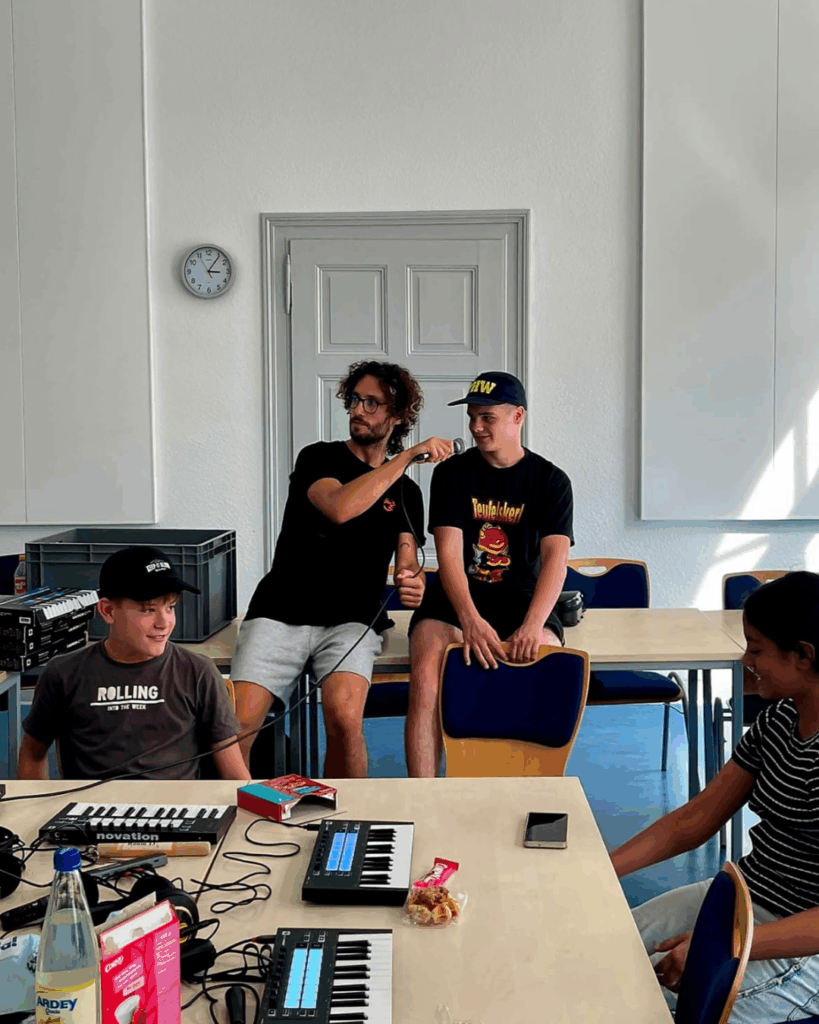 Der Workshop-Leiter hält einem Jungen ein Mikrofon hin. Zwei weitere Jugendliche sitzen an einem Tisch mit zwei Keyboards und Snacks.